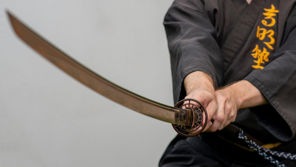 Ein Katana (Samuraischwert) wird in der Chudan no Kamae-Position gehalten. Der Schwertkämpfer trägt eine schwarze Trainingskleidung mit goldenen Schriftzeichen.